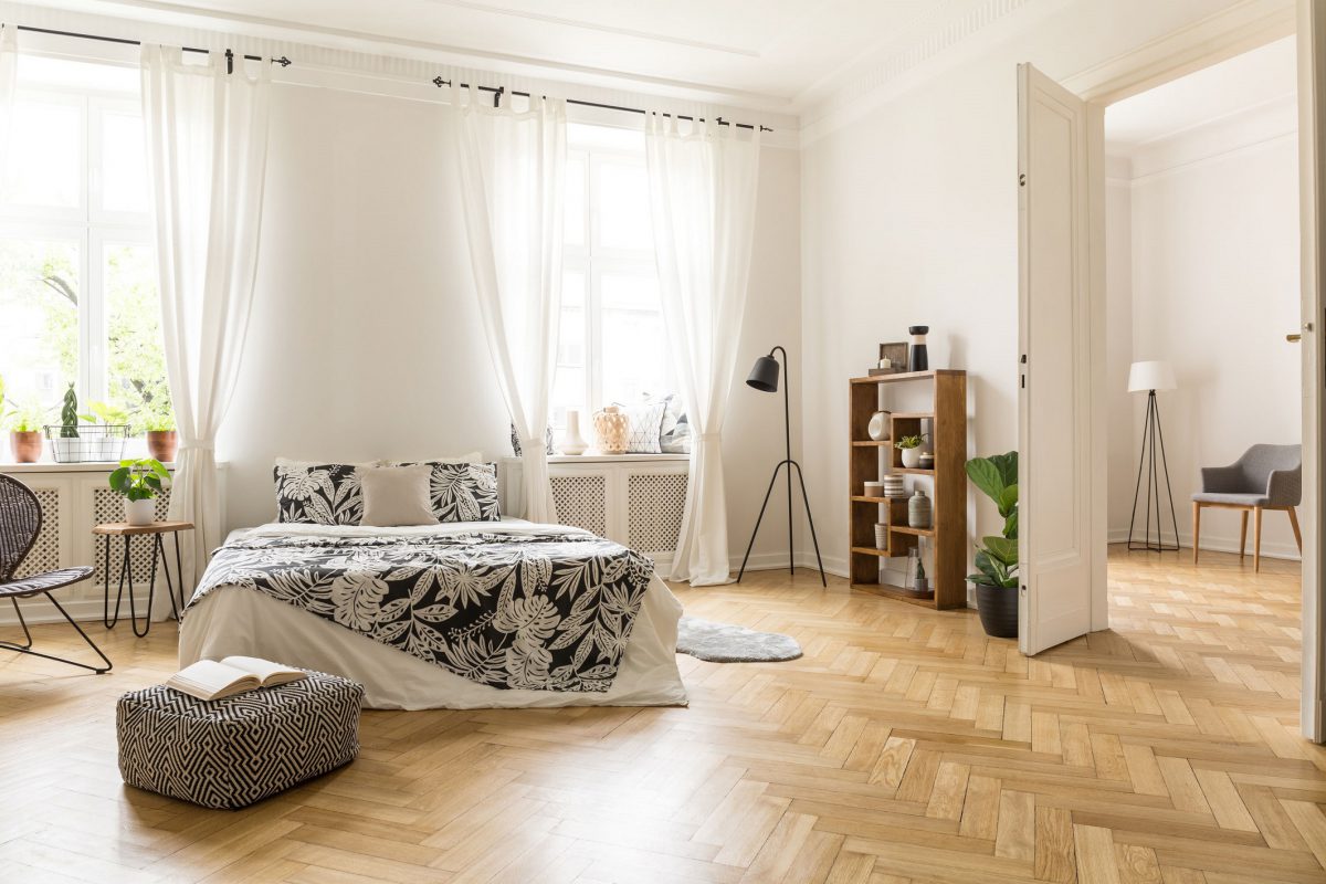 View at two rooms with white walls and herringbone parquet. A bed, a pouf, lamp bookcase and armchair in a natural style apartment interior. Real photo.