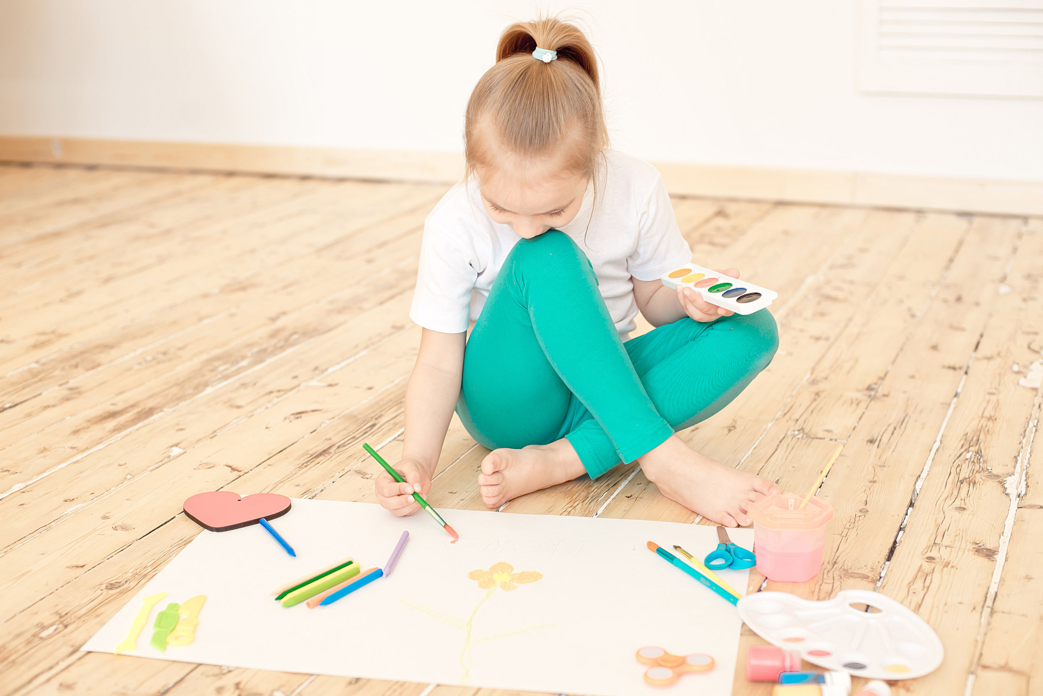 Little blonde girl paints on big white paper sitting on the floor indoors.