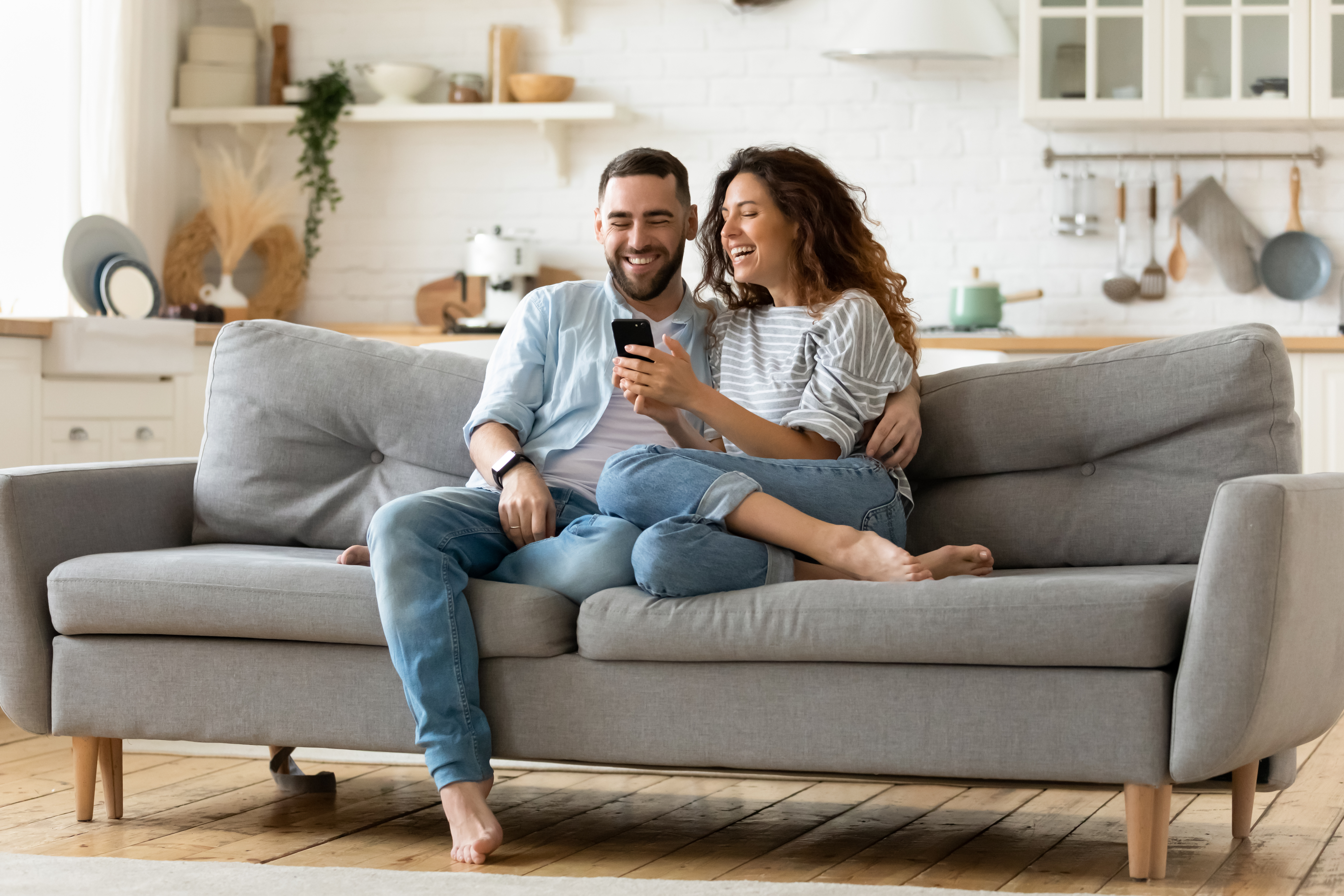 Happy young woman and man hugging, using smartphone together, sitting on cozy couch at home, smiling overjoyed wife and husband looking at phone screen, sitting on sofa in modern living room